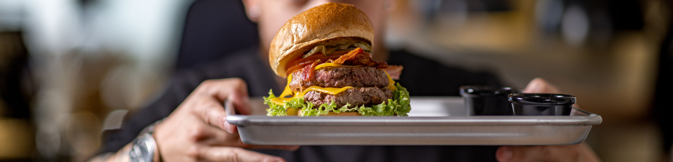Burger auf Tablett mit Salat, Patty, Käse, Bacon, Gurken und Bun, gehalten von einer Person, im Hintergrund unscharf. Neben dem Burger stehen zwei schwarze Saucenschalen.