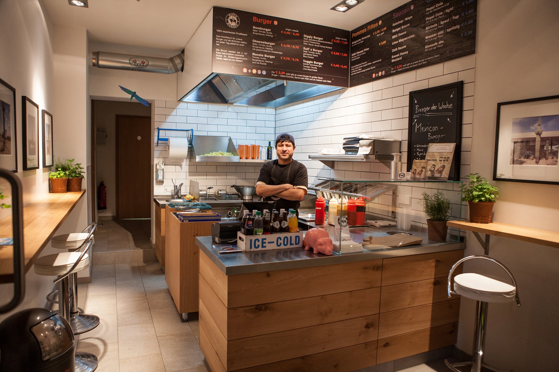 Innenansicht von einem "Ruff's Burger" mit Theke, Küche im Hintergrund, Sitzmöglichkeiten, weißen Fliesen und Menütafeln. Hinter der Theke steht ein Mitarbeiter.