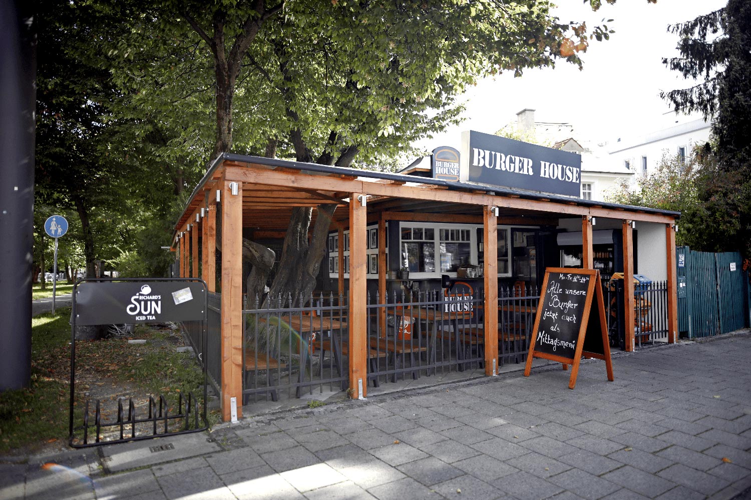 Außenansicht des Burgerrestaurants „Burger House“ mit überdachter Terrasse, Holzkonstruktion und einem großen Aufsteller mit Tagesangebot auf dem Gehweg.
