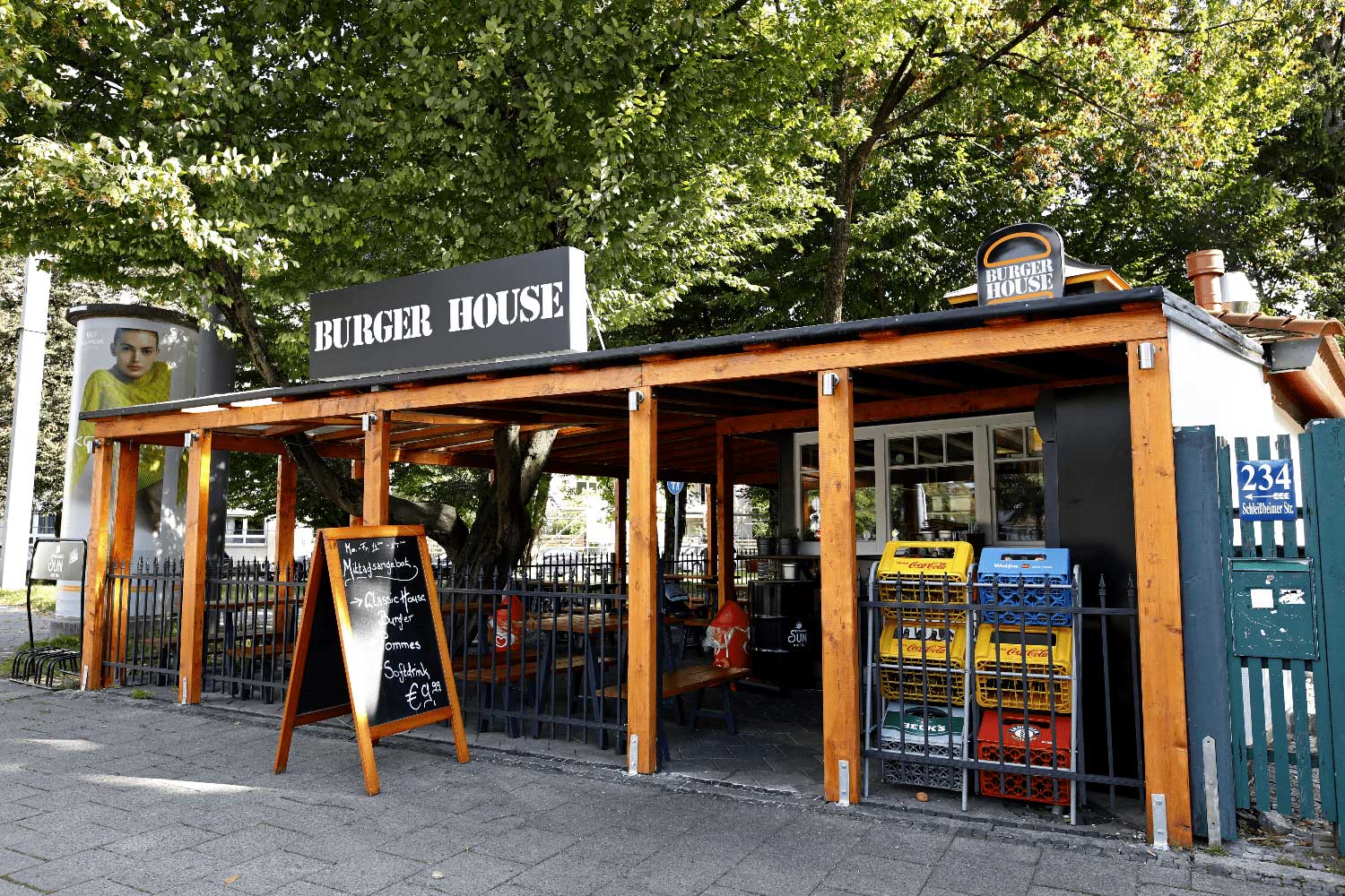 Frontansicht des „Burger House“ mit überdachter Sitzfläche, Getränkekisten am Eingang und einem Werbeschild mit Menüangebot auf dem Bürgersteig.