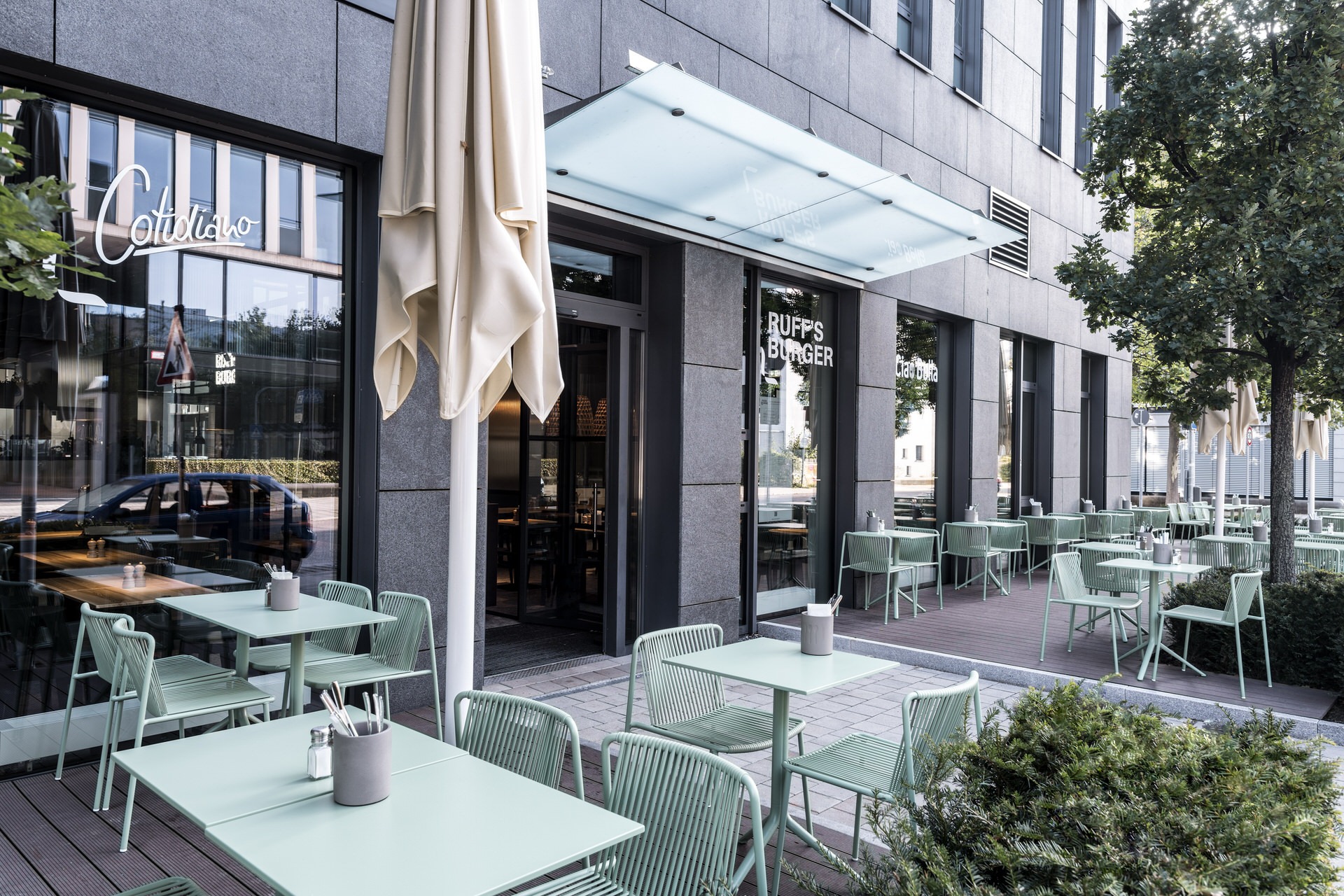 Außenansicht von "Ruff's Burger" und "Cotidiano" mit Terrasse, hellgrünen Tischen und Stühlen, Sonnenschirmen und Glasdach über der Eingangstür.