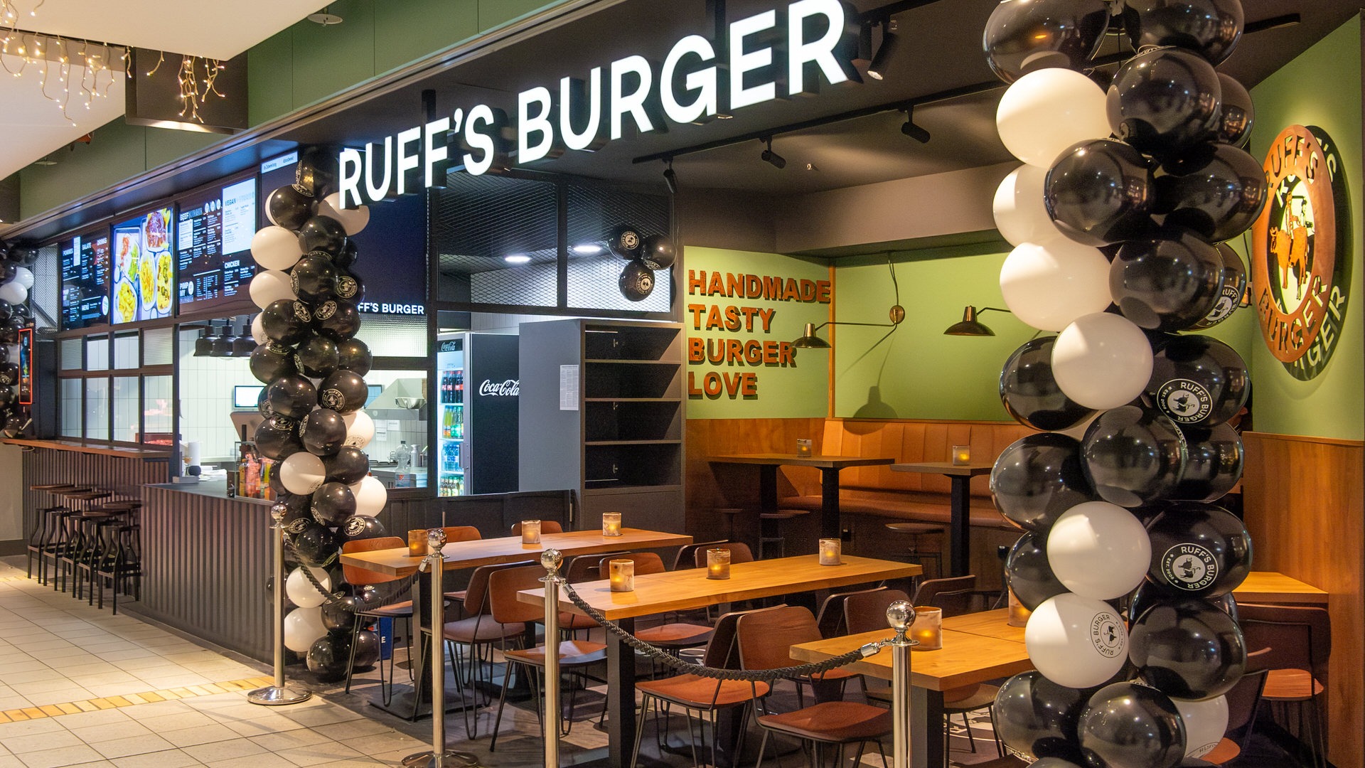 Außenansicht von einem "Ruff's Burger" Imbiss mit Ballondekoration, Sitzbereich, grüner Wand und beleuchtetem Schriftzug.