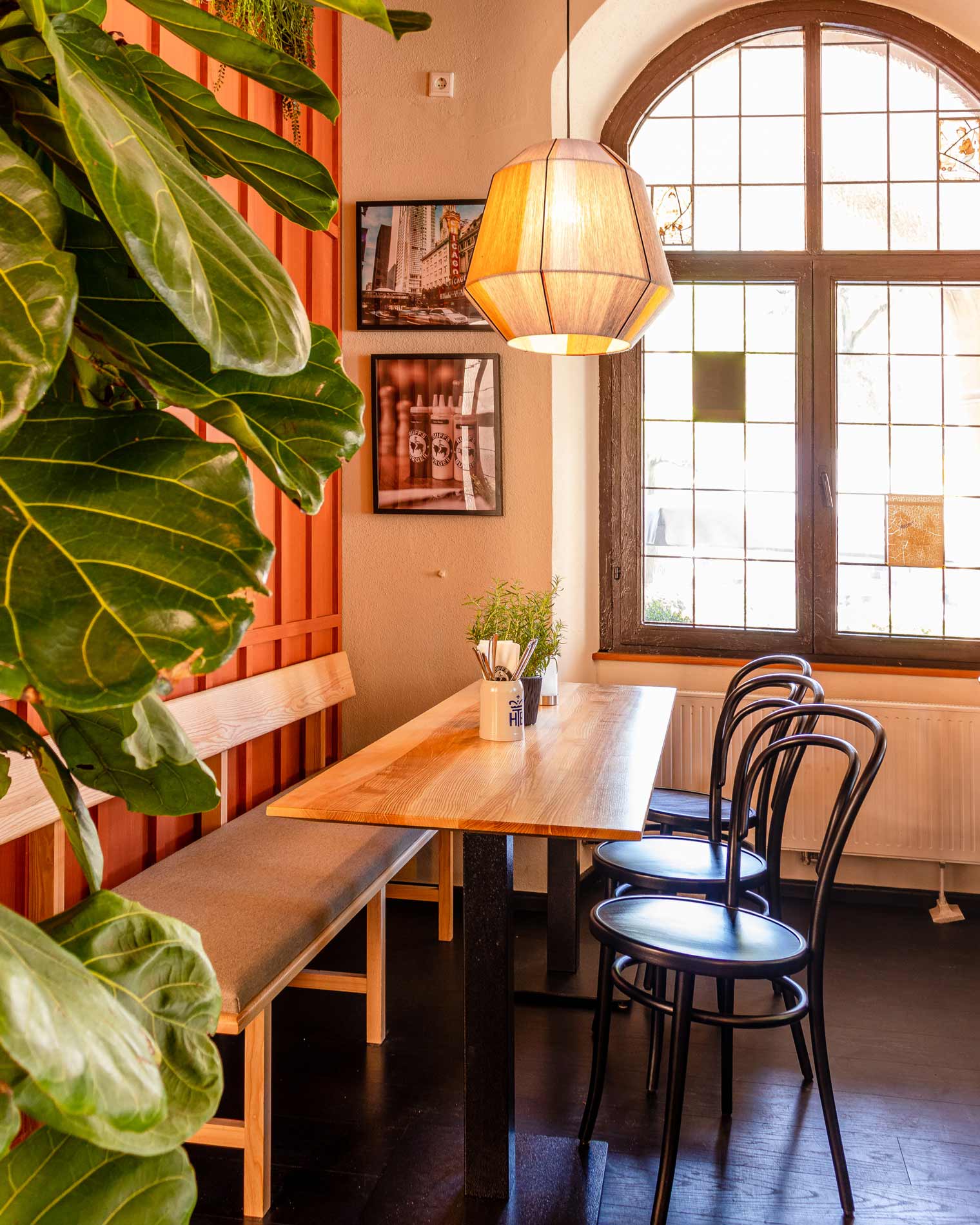 Detailansicht von einem "Ruff's Burger"-Sitzbereich mit großer Pflanze, Holztisch mit dunklen Holzstühlen und einer Sitzbank, Wandbildern, Holzfenster und Hängelampe.