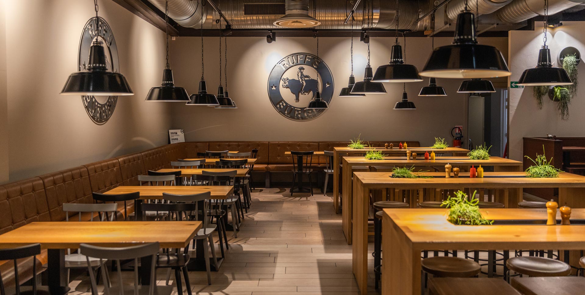 Innenansicht von "Ruff's Burger" mit Holztischen, hohen und niedrigen Sitzbereichen, brauner Polsterbank, schwarzen Hängelampen und großem Wandlogo.