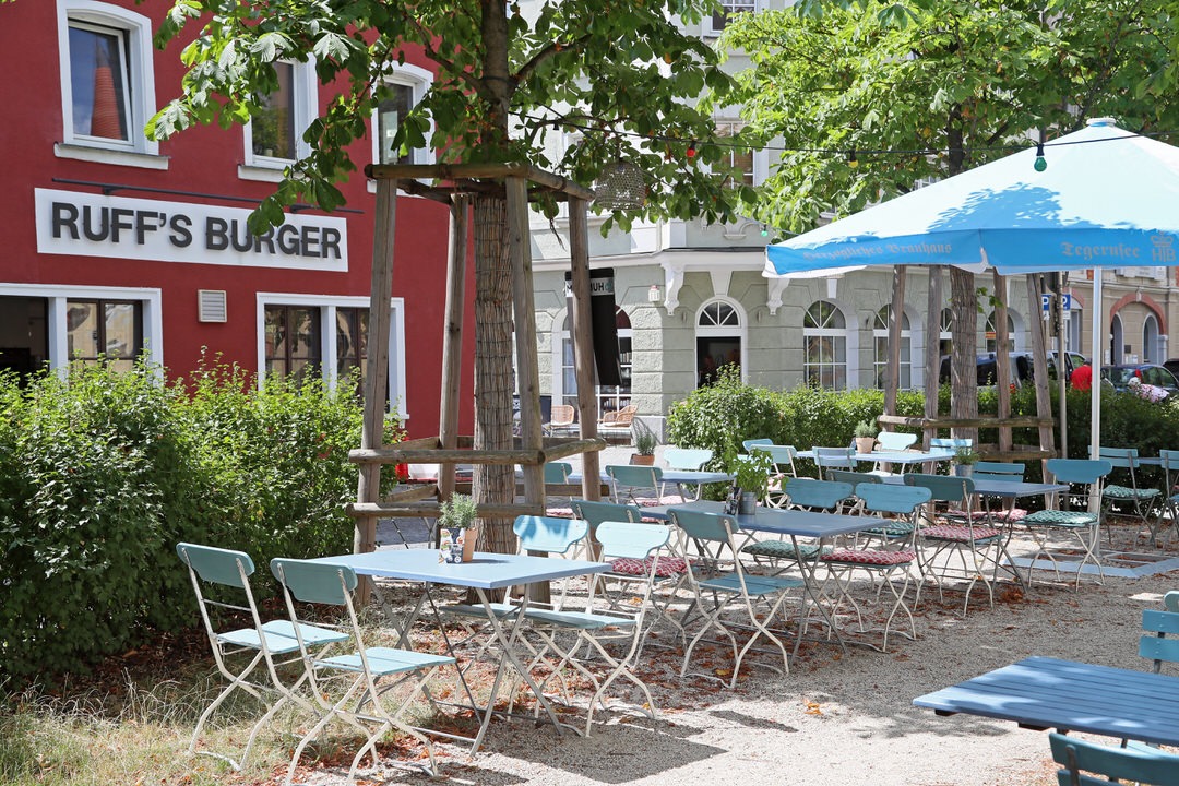 Außenbereich von "Ruff's Burger" mit blau-weißen Klapptischen, einem blauen Sonnenschirm, Bäumen und Blick auf die rote Restaurantfassade.