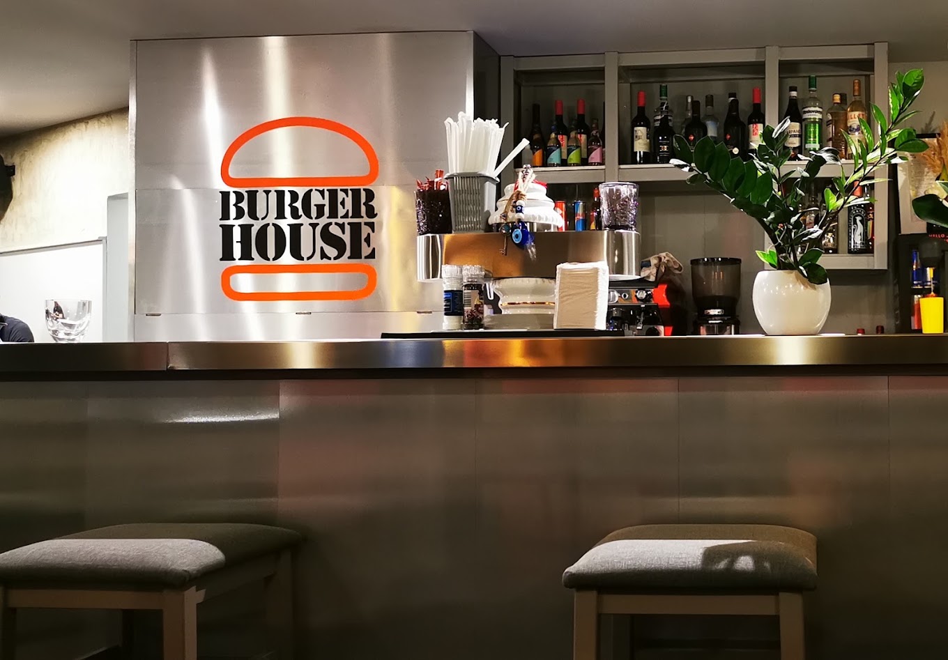 Moderner Bartresen mit beleuchtetem „Burger House“-Logo, Barhockern, Getränkeflaschen und Pflanze auf dem Tresen.