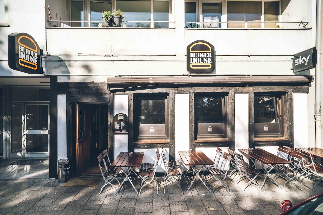 Außenansicht des Burger House mit mehreren Holztischen und Metallstühlen auf dem Gehweg.