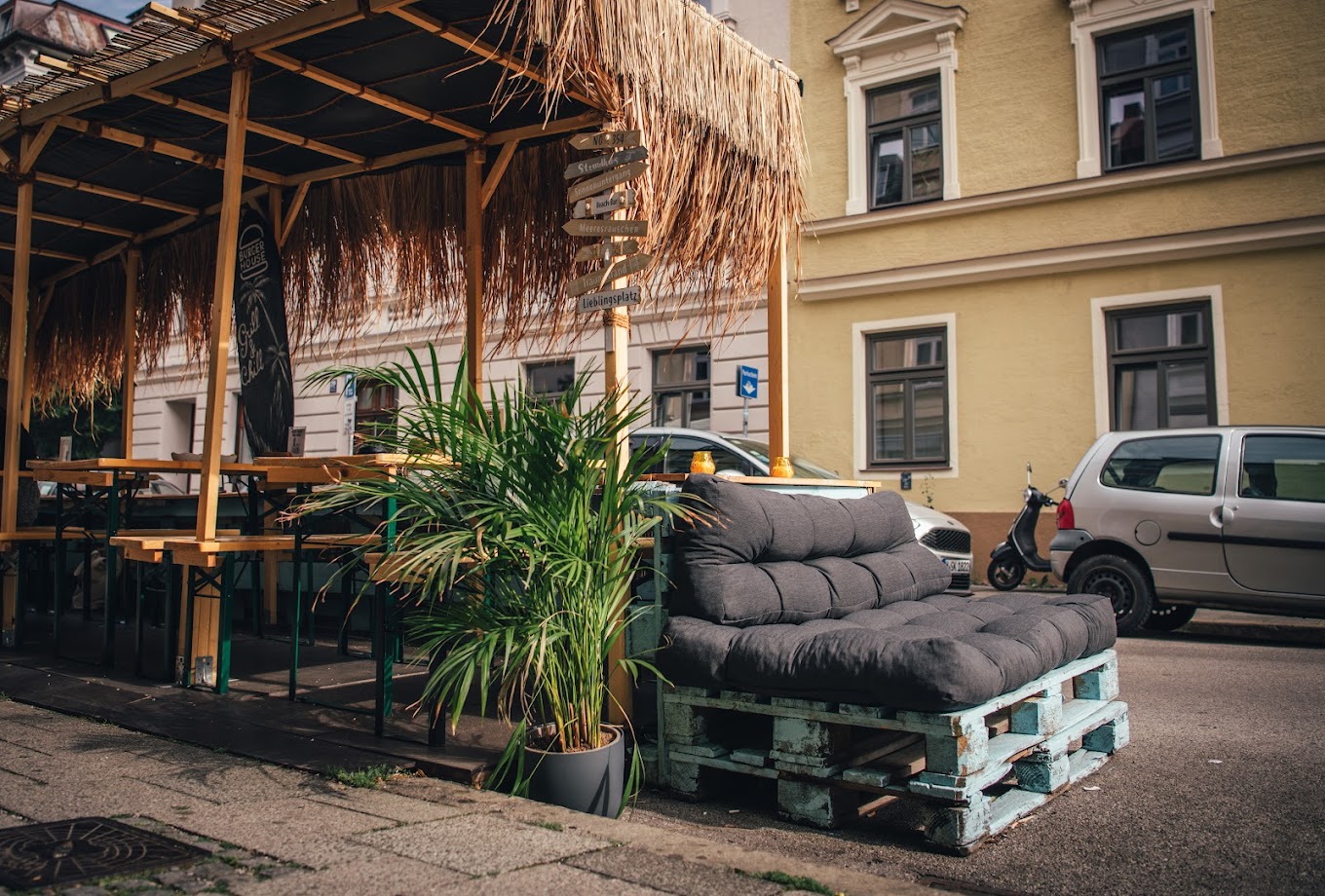 Außensitzbereich von "Burger House" mit Strohdach, Paletten-Sofa, Pflanzen und Blick auf die Straße.