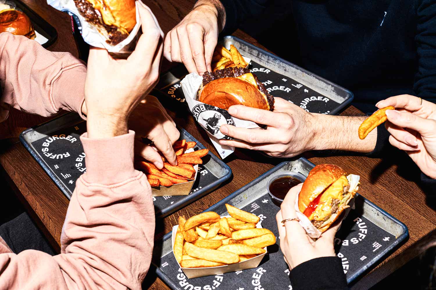Drei Personen essen Burger und Pommes an einem Tisch, Tabletts mit "Ruff's Burger" Branding und Dipschalen.