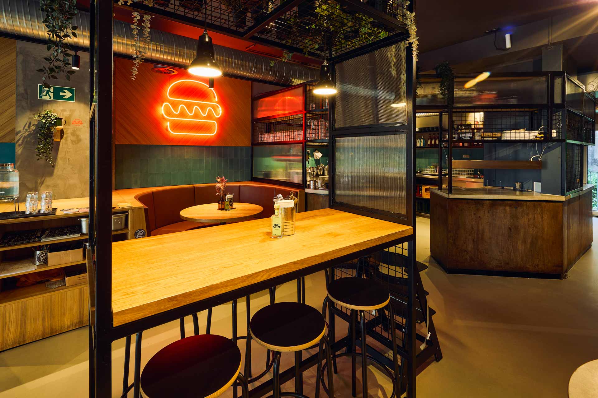 Innenansicht von "Ruff's Burger" mit runder Sitzbank, Holztischen, Neon-Burger-Symbol, grünen Wandfliesen. Gelber Gitter-Hochtisch mit Barhockern.