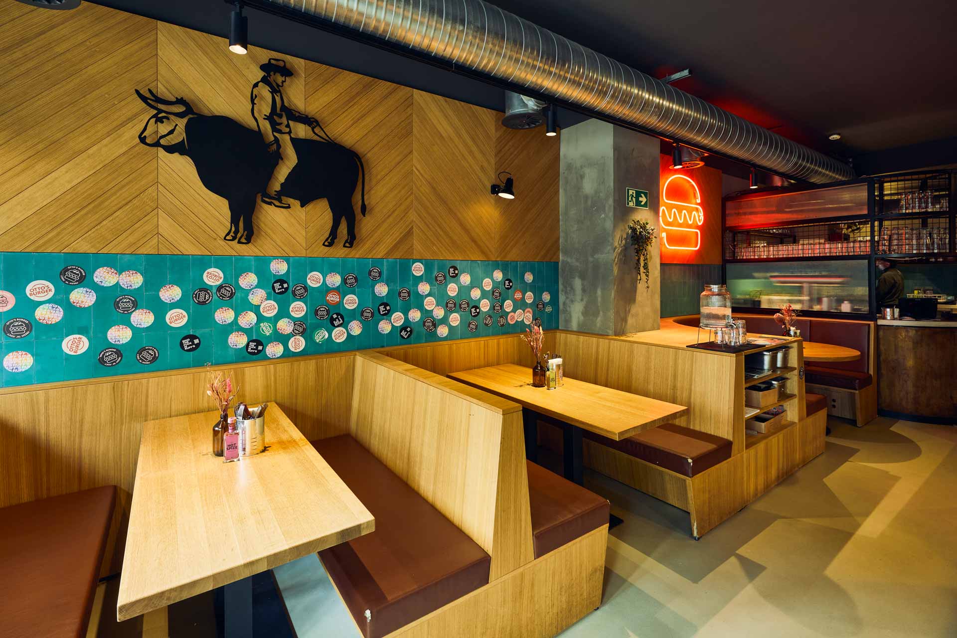 Innenansicht von "Otto's Burger" mit Holztischen, braunen Sitzbänken, grünen Wandfliesen und diagonalen Holzpaneelen.